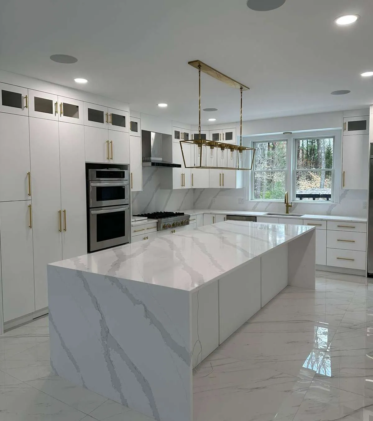 White flat-front kitchen with brass hardware and a marble waterfall island, cabinetry by Quality Design Cabinet